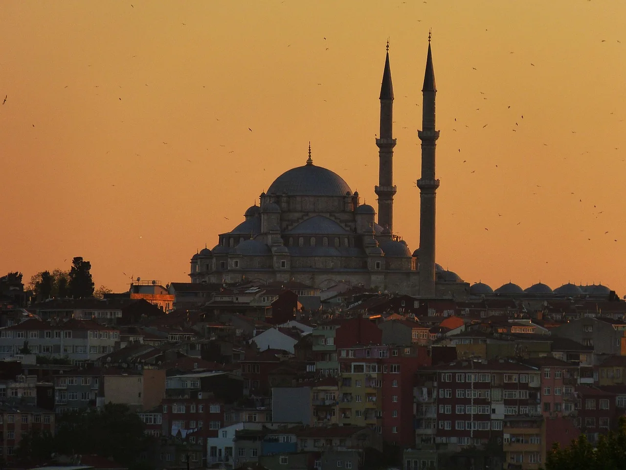 A domed mosque with two minarets above hillside houses in orange evening light