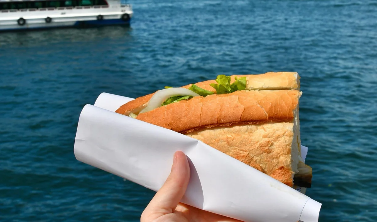 Balık ekmek sandwich held beside the waterfront, with a ferry in the background