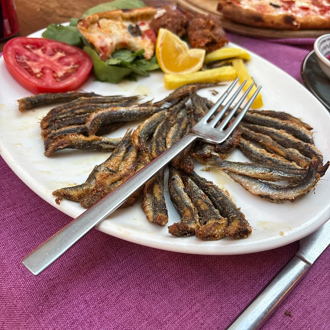 Fried anchovies arranged on a white plate with a fork, lemon wedges and sliced tomato