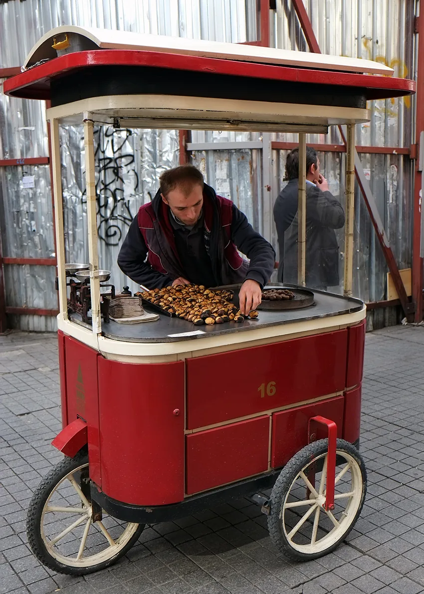 A vendor arranging roasted chestnuts on a red street cart in Istanbul